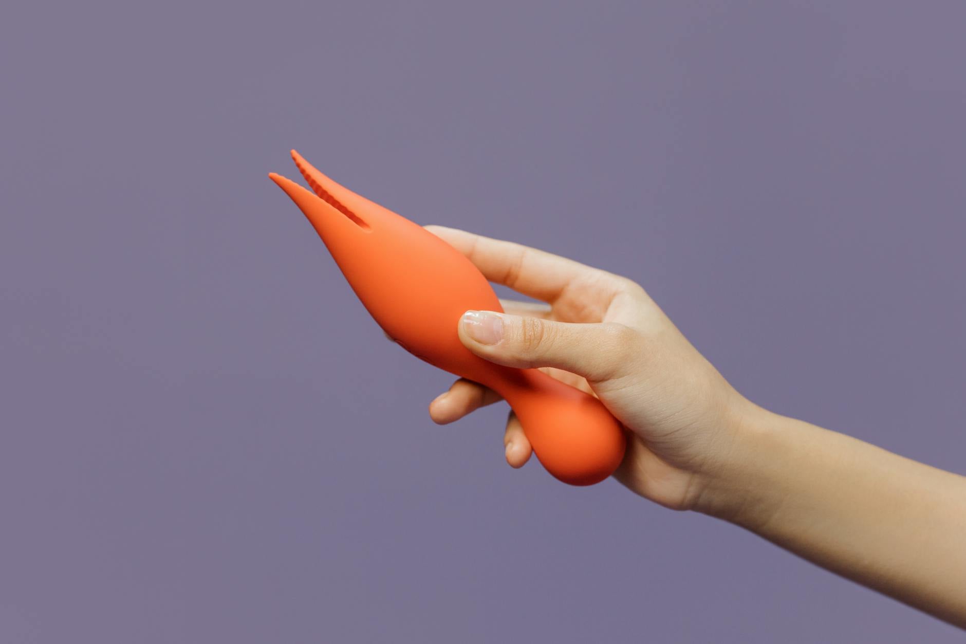 A close-up of a hand holding an orange vibrator against a minimalistic purple backdrop, showcasing modern sensuality.